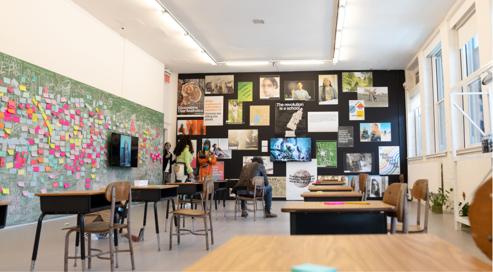 The Slow Factory exhibit is filled with school desks, a chalkboard wall covered in words and post-it notes, and a black wall in the back covered with posters.