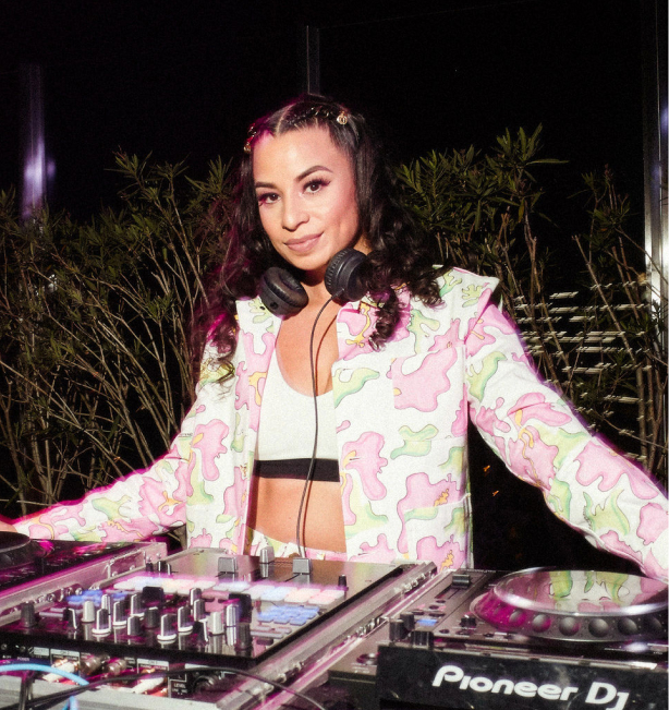 Jasmine Solano, a tan woman with long curly brown hair wearing a pink and green floral blazer. She stands behind her DJ equipment.