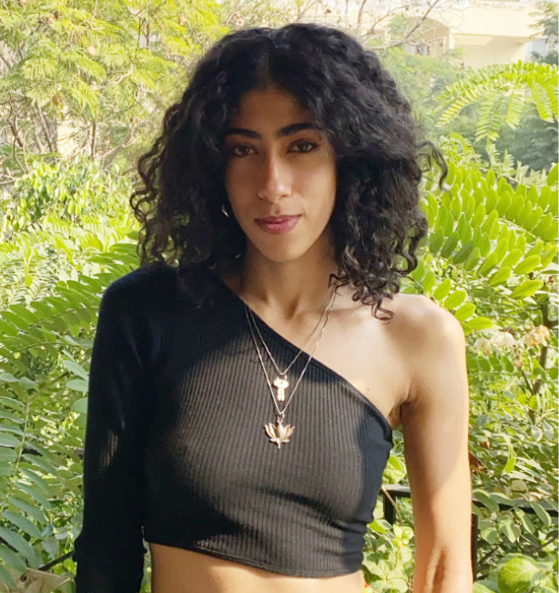 Felukah, an Egyptian woman, with curly black hair and tan skin wearing a black one-shoulder crop top and some gold necklaces. She stands in front of bright green foliage.