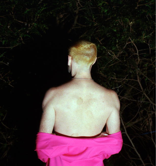 This image shows the back of Collis Browne, a person with short platinum hair, wearing a fuschia shirt that is draped down to the elbows to show off their shoulders. This person is standing in the darkness and a bright light is illuminating them.