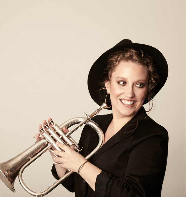 Rachel Thierrien, a white woman with strawberry blonde hair, wearing a suit jaclet and top hat. She is smiling a holding her trumpet while standing in front of a beige background.