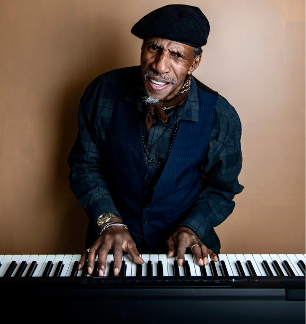 Sam Waymon, a older black man wearing a black beret, a dark green plaid shirt, and navy vest. She looks into the camera as he plays the piano.
