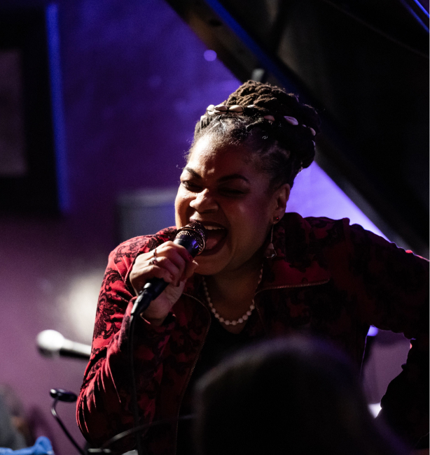 Phyllis Kee, a black woman with braided hair that sits in a ponytail. She is wearing a red jacket and sings into her microphone.