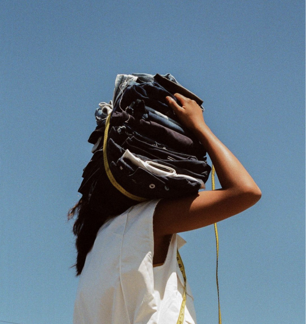 AAn image Krista Sison, the person behind Multitudes Studio. Her face is covered because she is holding a stack of old denim scraps on her shoulder. She is wearing a white top that pops against the blue sky.