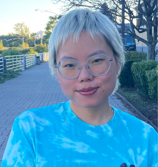 Mae is an Asian woman with short plantinum hair and big circular glasses. She is standing outdoors and is wearing a bright blue tie dyed t-shirt.