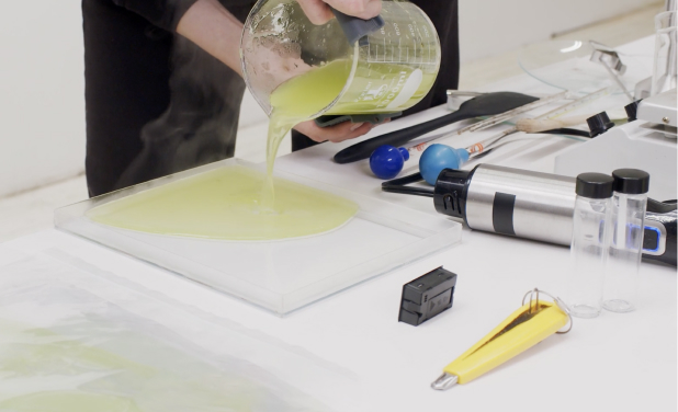 A close up image of a desk in a material science lab, where a person is pouring a  soft lime-colored liquid into a plastic tray. Other lab equipment is scattered on the desktop.