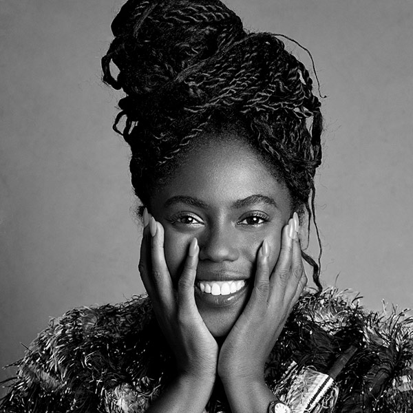 Abrima Erwiah, A black and white image of a smiling Black woman with braided hair, tied up. Her chin is rested on both her hands.