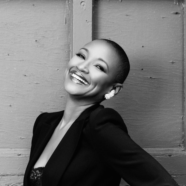 Christine Platt, A black and white image of a Black woman sporting a shaved head and dark blazer. Her head is title back as she smiles.